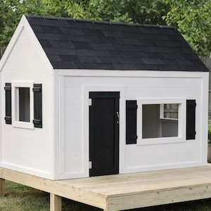 Peut inclure: Une cabane blanche avec un toit en bardeaux noirs, des volets noirs et une porte noire. La cabane est posée sur une terrasse en bois et possède deux fenêtres. La cabane est dans un jardin.