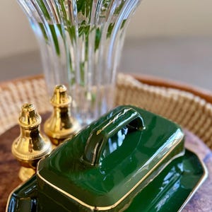 May include: A glossy dark green ceramic butter dish with a matching lid and a gold-toned trim along the edges. The rectangular lid features a central handle, and the base has a wide rim. Two small, polished gold-toned metal salt and pepper shakers.