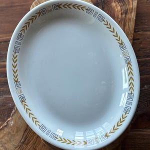 May include: White oval serving platter with a gold and black geometric and leaf design around the rim. The platter is empty and sits on a wooden surface. A vintage piece for serving food.
