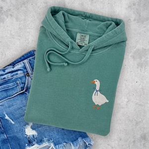 A green hoodie with a white goose embroidered on it, along with a pair of blue jeans, are laid out on a concrete surface.