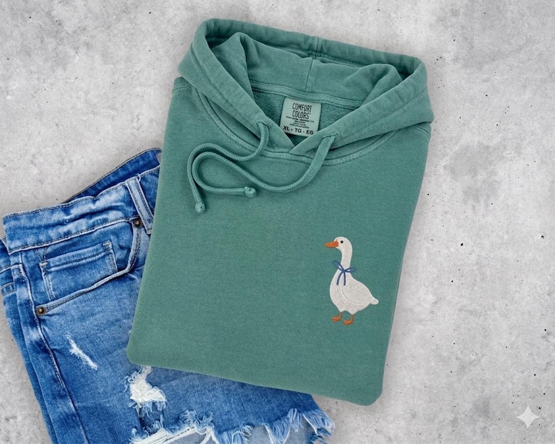 A green hoodie with a white goose embroidered on it, along with a pair of blue jeans, are laid out on a concrete surface.