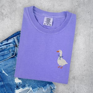 a purple t-shirt with a white duck embroidered on it, placed on top of a pair of blue jeans with frayed edges.