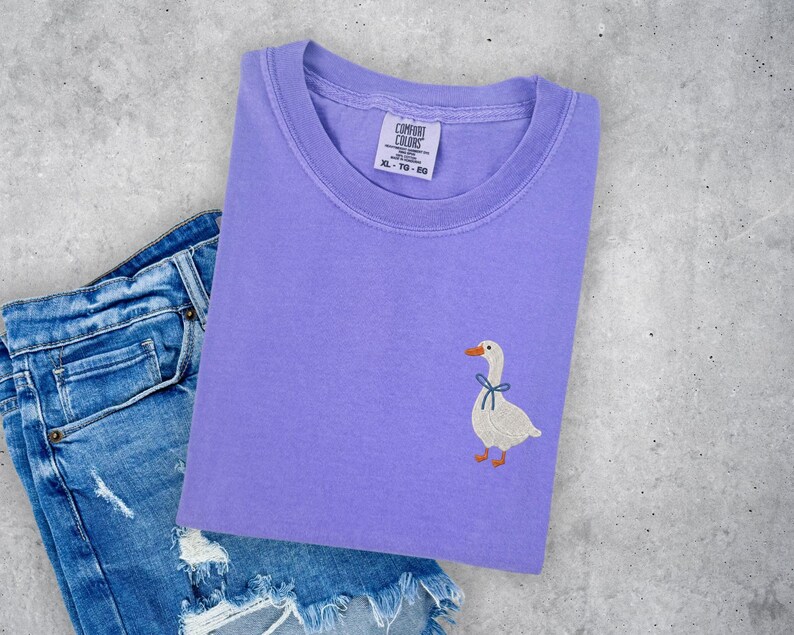 a purple t-shirt with a white duck embroidered on it, placed on top of a pair of blue jeans with frayed edges.