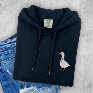 A black hoodie with a white duck embroidered on the front, placed on top of a pair of blue jeans.