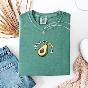 May include: A teal green t-shirt with the words "AVOCADO CLUB" and an avocado graphic. The shirt is folded and displayed with blue jeans, white slip-on shoes, and a silver necklace. A dried palm leaf is also in the image.