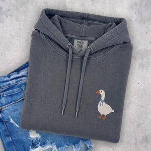 A gray hoodie with a white duck embroidered on it, along with a pair of blue jeans, are laid out on a concrete surface.
