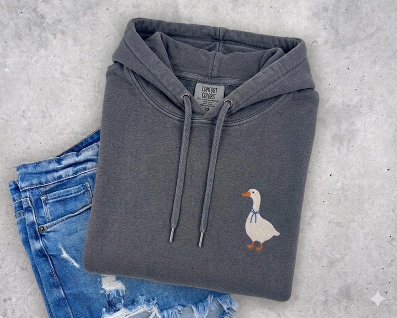 A gray hoodie with a white duck embroidered on it, along with a pair of blue jeans, are laid out on a concrete surface.