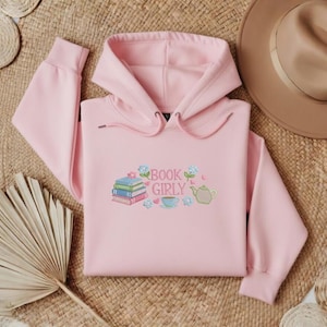 May include: A light pink hoodie with the embroidered text "BOOK GIRLY" and illustrations of books, flowers, a teapot, and a teacup. The hoodie is laid on a woven mat with a brown hat and decorative woven coasters.