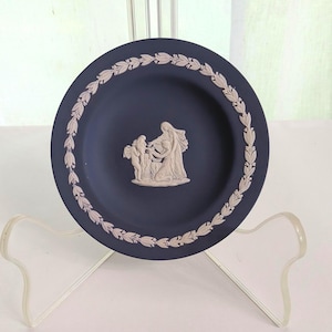 May include: A navy blue decorative plate with a white relief design. The plate features a central relief of figures and a white leaf border. The plate is displayed on a clear plastic stand.