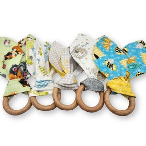 May include: Five baby teething rings with wooden rings and fabric ears. The fabric ears feature various patterns, including animals, bees, and geometric designs. The colors include green, blue, yellow, and gray.