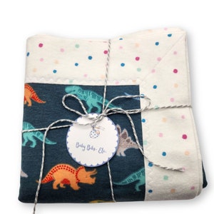 May include: A blue and white baby blanket with a dinosaur print. The blanket is tied with twine and has a tag that says "Baby Bibs, Etc."