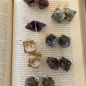 Pode incluir: Uma coleção de brincos de resina feitos à mão em várias cores e designs, incluindo formas de dados e inclusões florais. Os brincos têm acessórios de metal dourado e prateado. Os brincos são exibidos em um livro aberto.