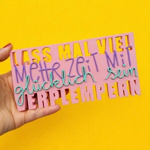 Könnte beinhalten: Ein rosa rechteckiges Schild mit ausgeschnittenen Buchstaben vor gelbem Hintergrund. Das Schild zeigt den deutschen Satz "Lass mal viel mehr Zeit Mit glücklich sein VERPLEMPERN" in Lila und Türkis.