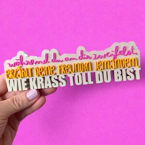 Könnte beinhalten: Ein weißes, mehrschichtiges Schild mit deutschem Text in Pink, Gelb und Weiß. Der Text lautet: "während du an dir zweifelst. Erzählt deine Freundin jemandem WIE KRASS TOLL DU BIST."