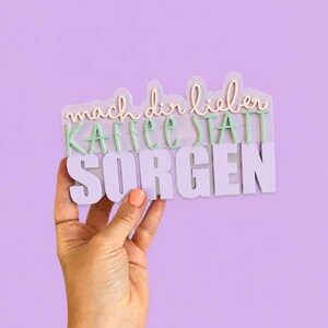 Könnte beinhalten: Ein mehrschichtiges Schild mit dem deutschen Text "mach dir lieber KAFFEE STATT SORGEN" in einer Kombination aus Schreib- und Blockschrift. Das Schild verwendet eine Farbpalette aus Helllila, zartem Rosa und Mintgrün mit erhabenen Buchstaben.