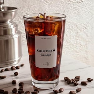 Op de afbeelding: Een glazen kaars gevuld met donkerbruine was, die op ijskoffie lijkt, met ijsblokjes. Het etiket luidt "COLD BREW Candle". Koffiebonen zijn rond het glas verspreid en een zilveren koffiemolen staat op de achtergrond.