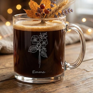 Puede incluir: Una taza de vidrio transparente llena de líquido oscuro, posiblemente café, adornada con un diseño floral y el nombre "Emma". La taza está coronada con un arreglo decorativo de follaje otoñal, incluyendo una hoja de arce, bayas y lavanda.