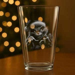 Puede incluir: Un vaso de pinta transparente con una ilustración de dibujos animados de una criatura verde y gruñona. El personaje tiene una expresión severa y se encuentra sobre un fondo borroso de un árbol de Navidad con luces doradas. El vaso está sobre una superficie de madera.