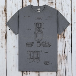 Puede incluir: Camiseta gris con un diseño de patente de micrófono vintage. El gráfico incluye diagramas detallados, fechas e información del inventor. Una prenda única para los amantes de la música y la tecnología vintage.