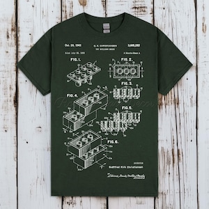May include: A dark green short-sleeve t-shirt featuring a white patent illustration of a toy building brick. The graphic includes technical drawings labeled Fig. 1 through Fig. 6, along with text detailing the patent date Oct. 24, 1961, and inventor.