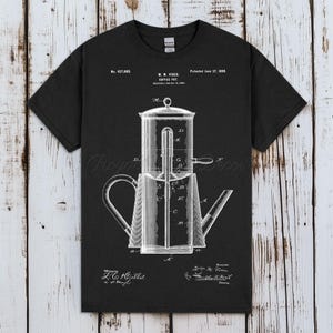 Puede incluir: Camiseta negra con un diagrama blanco detallado de una cafetera, incluyendo etiquetas e información de patente. El diseño muestra los componentes internos y la estructura de la cafetera. La camiseta tiene un cuello redondo clásico.