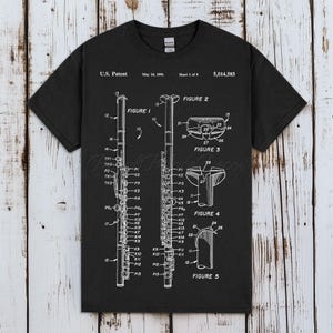以下が含まれることがあります： 白のフルートの図が描かれた黒いTシャツ。ラベルと図が含まれています。シャツには「U.S. Patent May 14, 1991 Sheet 1 of 4 5,014,585」と書かれています。デザインは楽器のコンポーネントの詳細なイラストです。