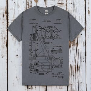 Op de afbeelding: Grijze t-shirt met een zwart-wit diagram van een drumstel, met tekst als "July 7, 1959" en "F. J. Ippolito Practice Drum Set." Het ontwerp lijkt op een patenttekening. Het shirt is gemaakt van zacht materiaal.