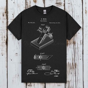 Pode incluir: Uma t-shirt preta de manga curta com uma impressão gráfica branca de uma ilustração de patente para um relógio de sol de G. Mehr, datada de 22 de setembro de 1874. O design inclui diagramas técnicos rotulados Fig 1, Fig 2 e Fig 3.