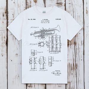 May include: White t-shirt featuring a black and white diagram of a musical instrument, a trumpet, with the text "H. Selmer Musical Instrument" and the date "Nov. 28, 1939." The shirt has a crew neck and short sleeves.