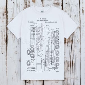 以下が含まれることがあります： 白のTシャツ。オーボエまたはイングリッシュホルンの詳細な白黒図。 「C.P. Müller」や「Patented Nov. 6, 1888」などのテキストが含まれています。 ヴィンテージの特許イラストです。