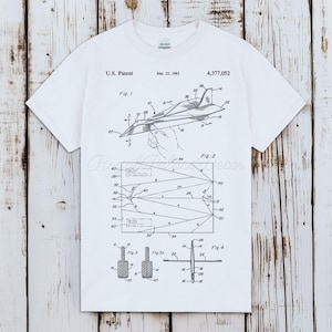 May include: A white short-sleeve t-shirt featuring a black graphic print of a U.S. Patent for a paper airplane, dated March 22, 1983, patent number 4,377,052. The design includes technical line drawings labeled Fig. 1, Fig. 2, Fig. 3, and Fig. 4.