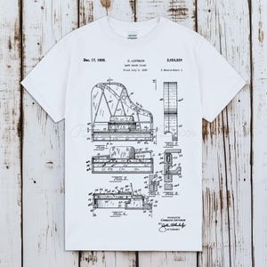 以下が含まれることがあります： 白のTシャツ。ベビーグランドピアノの白黒図面がプリントされています。図面には技術図面、ラベル、日付「1935年12月17日」が含まれています。シャツには発明者の名前と特許番号も含まれています。