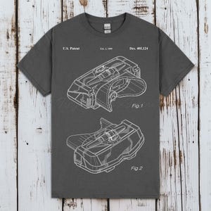 Pode incluir: Uma t-shirt cinzenta escura de manga curta com um gráfico de patente dos EUA em branco para o Des. 405,124 datado de 2 de fevereiro de 1999. A impressão exibe dois desenhos técnicos de linhas rotulados Fig. 1 e Fig. 2 de um dispositivo.