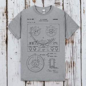 Puede incluir: Una camiseta gris de manga corta con una ilustración técnica negra impresa que representa la patente de una máquina de cinta magnética. El gráfico incluye diagramas etiquetados como Fig 1, Fig 2 y Fig 3, junto al texto Oct. 31, 1961, W. A. Ellmore, 3,006,650.