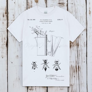 Könnte beinhalten: Ein weißes Kurzarm-T-Shirt mit einer schwarzen Patentzeichnung eines Bienenrauchers. Die Grafik enthält technische Diagramme, den Text Dec. 26, 1950, 2,535,177, Closure for Beehive Smokers sowie drei detaillierte Zeichnungen von Bienen.