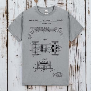 Könnte beinhalten: Ein graues Kurzarm-T-Shirt mit einer schwarzen technischen Patentzeichnung eines elektrischen Spielzeugautos und einer Fahrbahn. Die Grafik enthält den Text March 22, 1938, A. E. Cullen, 2,112,072 sowie detaillierte Diagramme mit Fig. 1, Fig. 2 und Fig. 3.