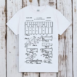 以下が含まれることがあります： 白のTシャツに、ピアノの鍵盤の特許の白黒図が描かれています。特許には日付、図、発明者の名前が含まれています。音楽愛好家のためのユニークなグラフィックTシャツです。