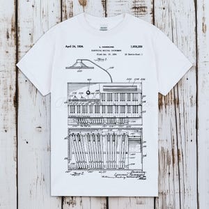 以下が含まれることがあります： 白のTシャツ。電気楽器の白黒図で、鍵盤とスピーカーがあり、「April 24, 1934」と「L. Hammond」の文字が書かれています。図は技術図面です。Tシャツは柔らかい素材で作られています。
