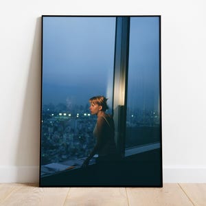 May include: A framed photograph of a woman looking out a large window at a city skyline at dusk. The woman is in silhouette, and the city lights are blurred in the background. The photograph is displayed on a white wall.