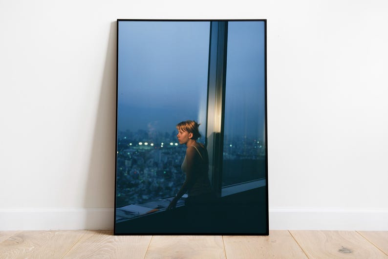 May include: A framed photograph of a woman looking out a large window at a city skyline at dusk. The woman is in silhouette, and the city lights are blurred in the background. The photograph is displayed on a white wall.