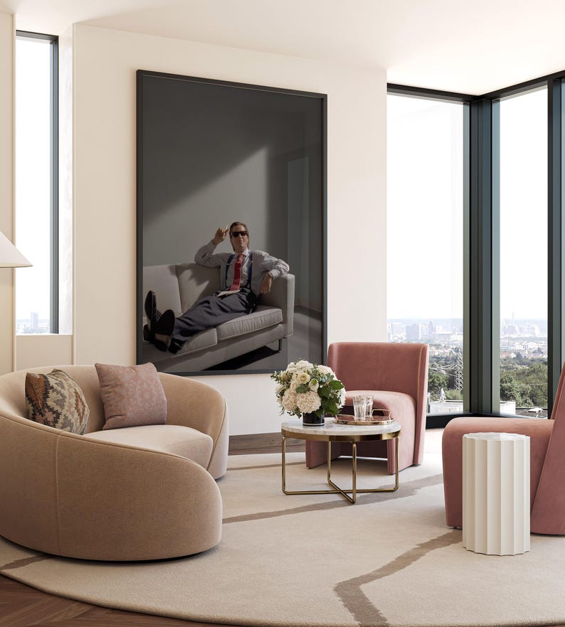 May include: A modern living room with a large framed artwork featuring a person on a sofa. The room includes two curved armchairs, a small round table, and a side table. The color palette is neutral with pink and beige accents.