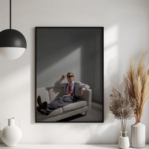 May include: Framed print featuring a man in a white shirt, red tie, and sunglasses, seated on a light-colored sofa. The image has a minimalist aesthetic with a dark background and a single light source.