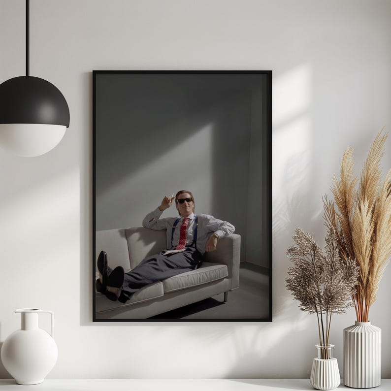 May include: Framed print featuring a man in a white shirt, red tie, and sunglasses, seated on a light-colored sofa. The image has a minimalist aesthetic with a dark background and a single light source.
