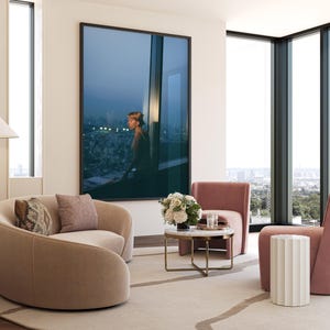 May include: A modern living room with a large framed artwork depicting a person looking out a window. Two curved beige armchairs, a pink armchair, and a small round marble table with gold legs are arranged on a light-colored rug. A white side table and large windows complete the scene.