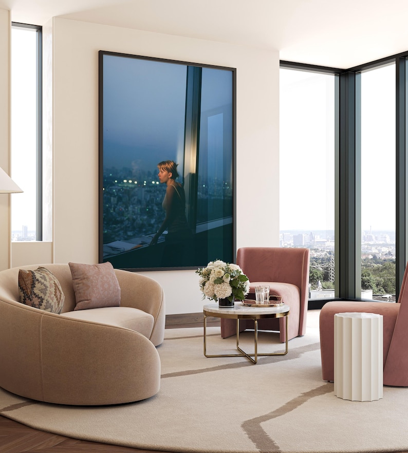 May include: A modern living room with a large framed artwork depicting a person looking out a window. Two curved beige armchairs, a pink armchair, and a small round marble table with gold legs are arranged on a light-colored rug. A white side table and large windows complete the scene.