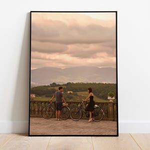 Op de afbeelding: Een ingelijste print met een stel met fietsen op een balkon met uitzicht op een bergketen. De lucht is bewolkt en de scène wordt verlicht door warm licht. De fietsen zijn zwart.
