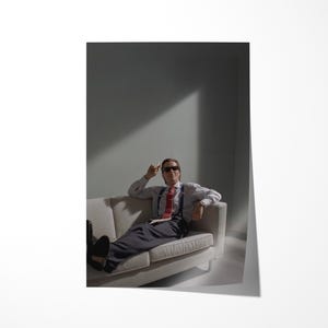 May include: A person wearing sunglasses, a white shirt, red tie, suspenders, and dark pants, reclines on a white sofa. The person is in a room with a gray wall and a beam of light.