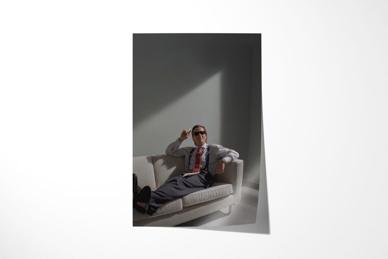 May include: A person wearing sunglasses, a white shirt, red tie, suspenders, and dark pants, reclines on a white sofa. The person is in a room with a gray wall and a beam of light.