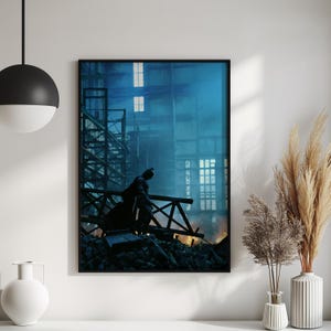 May include: A framed print featuring a dark figure in a cape, silhouetted against a blue-toned industrial backdrop. The figure stands on a wooden structure amidst rubble, with a cityscape visible in the background. The artwork is framed in black.