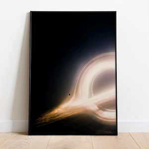 Pode incluir: Uma impressão artística emoldurada que retrata um buraco negro. A imagem apresenta um anel brilhante e brilhante de luz contra um fundo escuro. A obra de arte é exibida em um piso de madeira e encostada em uma parede branca.